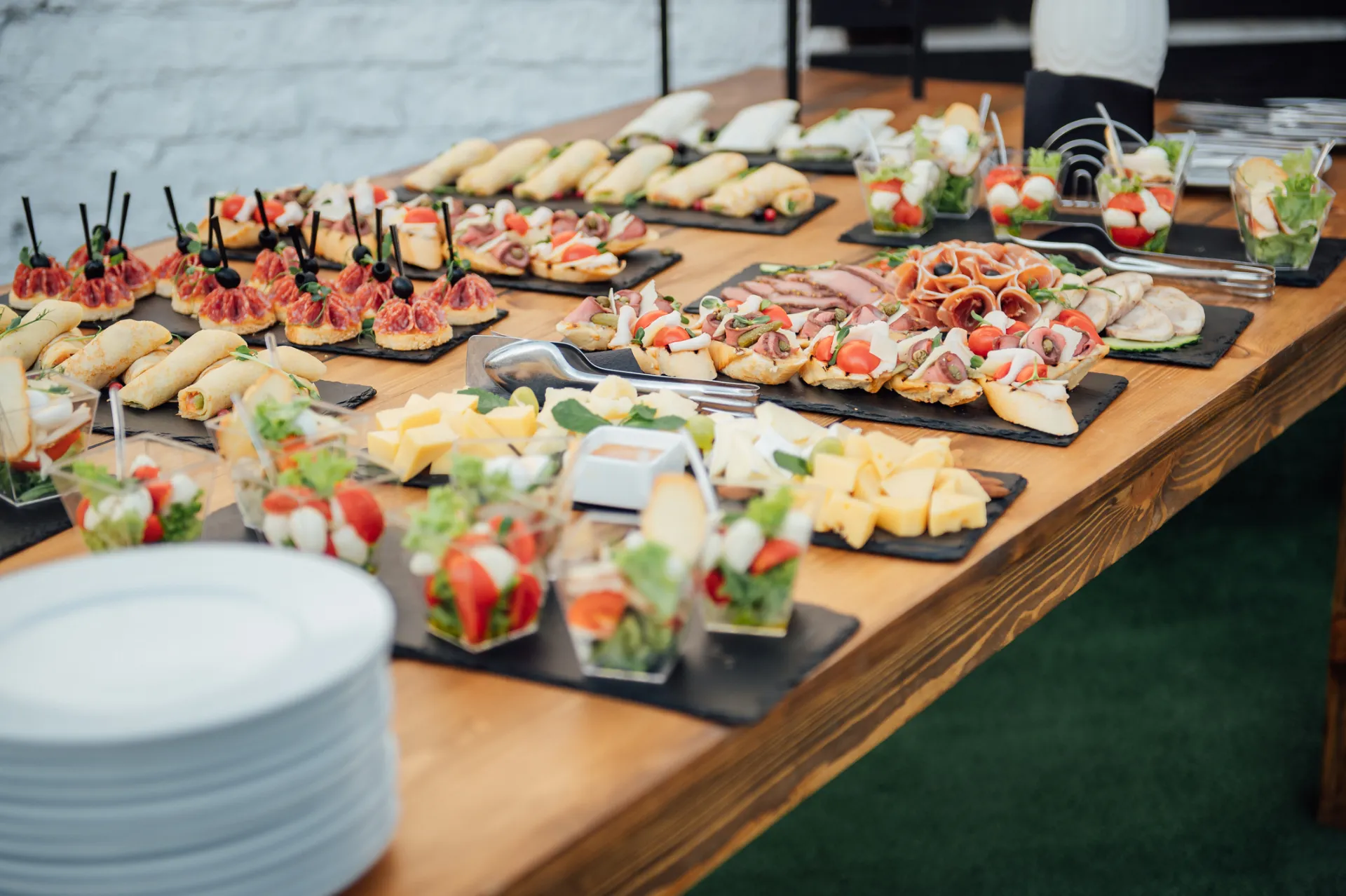 Catering-Buffet mit verschiedenen Fingerfood-Häppchen, Aufschnitt, Käse und Caprese-Salat in Gläsern auf Schieferplatten und einem Holztisch.